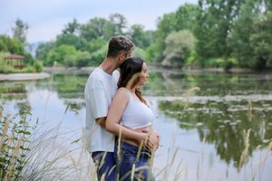 Couple standing by a serene lake in a lush park, woman cradling pregnant belly in a maternity photo shoot.