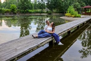 Couple sitting together on a wooden dock by a calm lake surrounded by lush green trees.