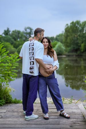 Couple on wooden dock by lake, pregnant woman in white top and jeans, man in white tee with ultrasound photos.