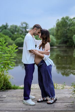 Couple sharing a tender moment on a wooden dock by a lake, woman visibly pregnant, both wearing white tops and jeans.