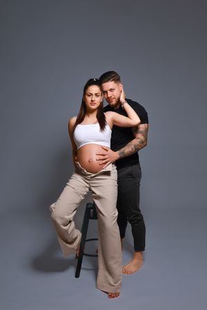 Expecting couple poses together during maternity photoshoot against gray backdrop.