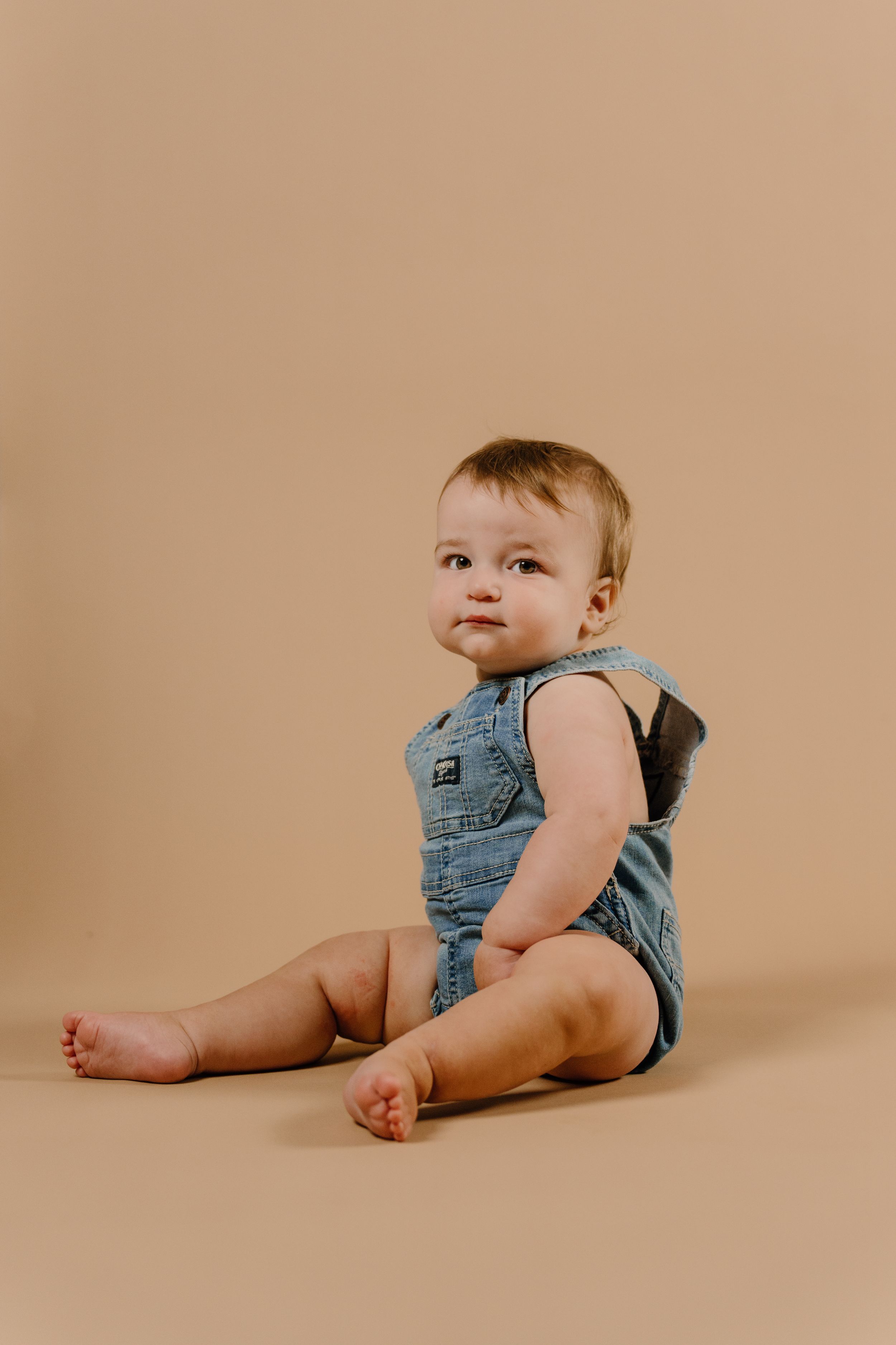 Baby wearing denim overalls sits on beige background in artistic studio portrait series showing different poses.