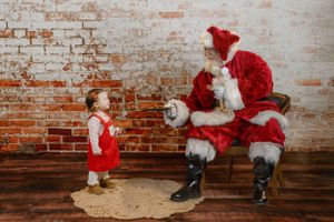 Santa Claus interacting with a small child in a red dress against a brick wall backdrop.