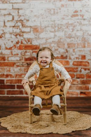 A small child wearing brown overalls sits cheerfully in a wooden chair on a macrame rug against a rustic brick wall.