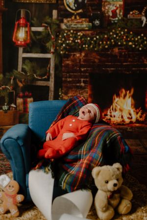 Cozy Christmas scene with red lantern, glowing fireplace, blue armchair, plaid blanket, and teddy bears by a decorated tree.
