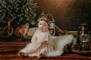 Professional studio photos of a baby in white dress and floral crown against dark moody holiday backdrop.
