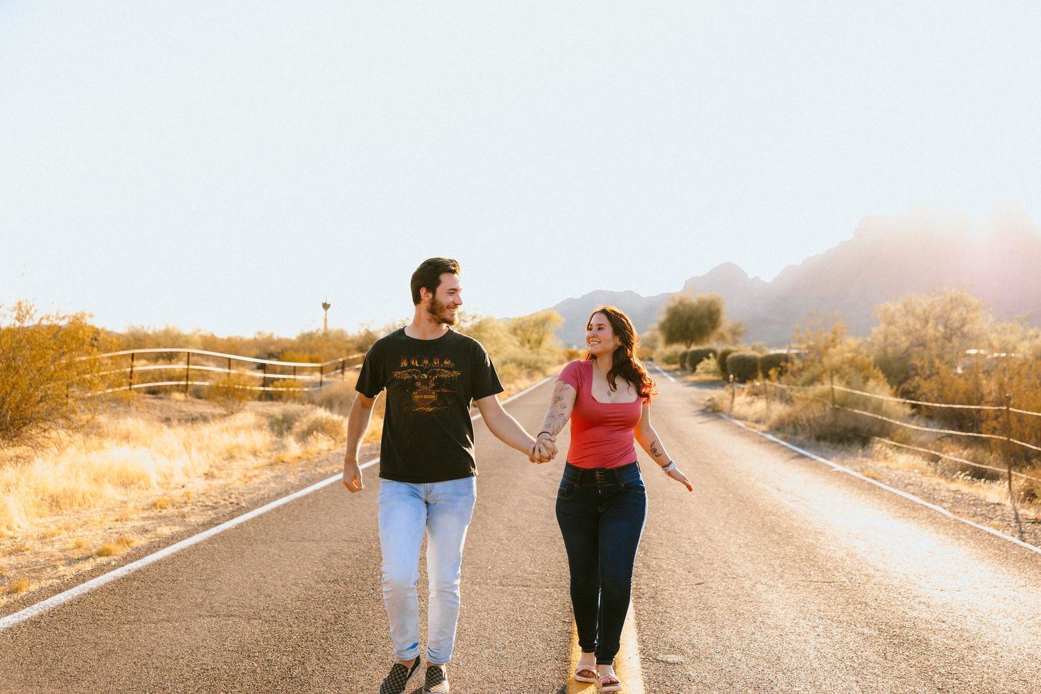 Two people walk together holding hands on desert road with mountains in background.