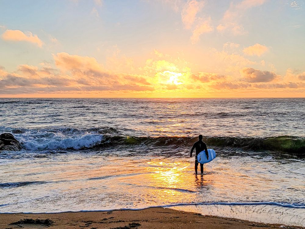 A surfer carries a white board into golden ocean waves at a stunning sunset beach.