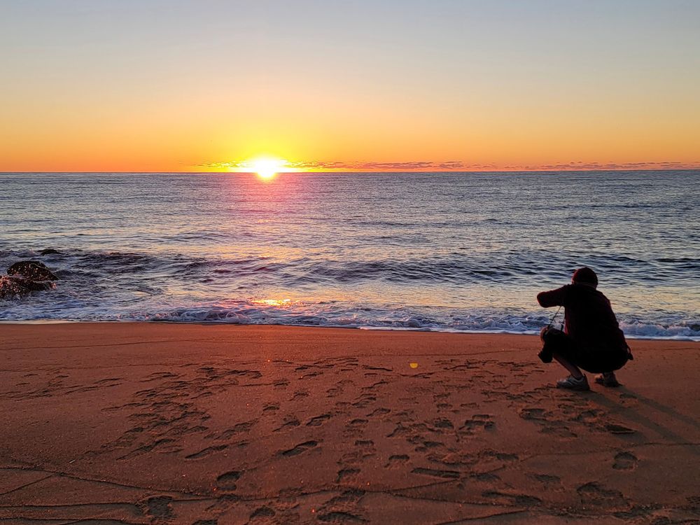 Person crouching on sandy beach photographing a vibrant golden sunset over the ocean waves.