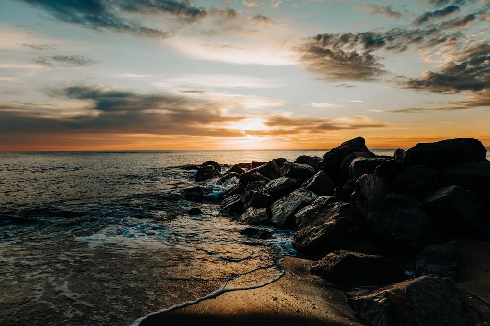 Rocky shoreline at sunset with golden sky, dramatic clouds, and gentle ocean waves washing over dark stones on a sandy beach.