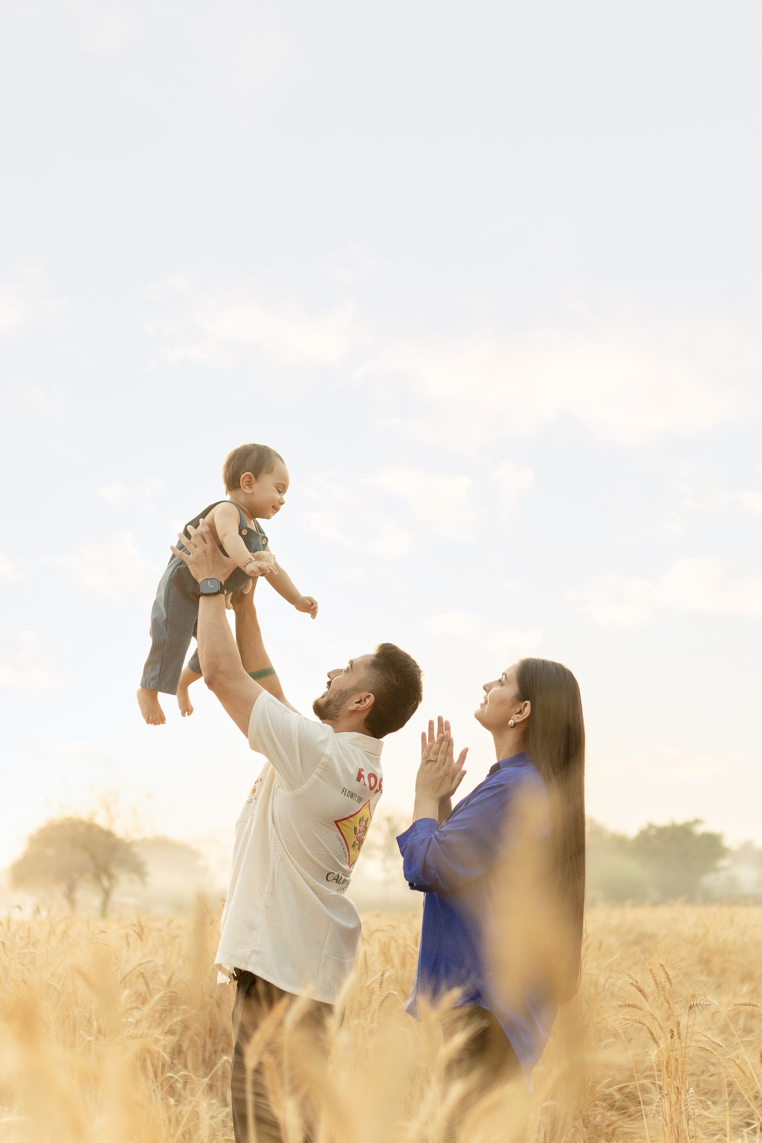 A joyful family moment captured in a golden wheat field at sunset as a baby is lifted into the air.