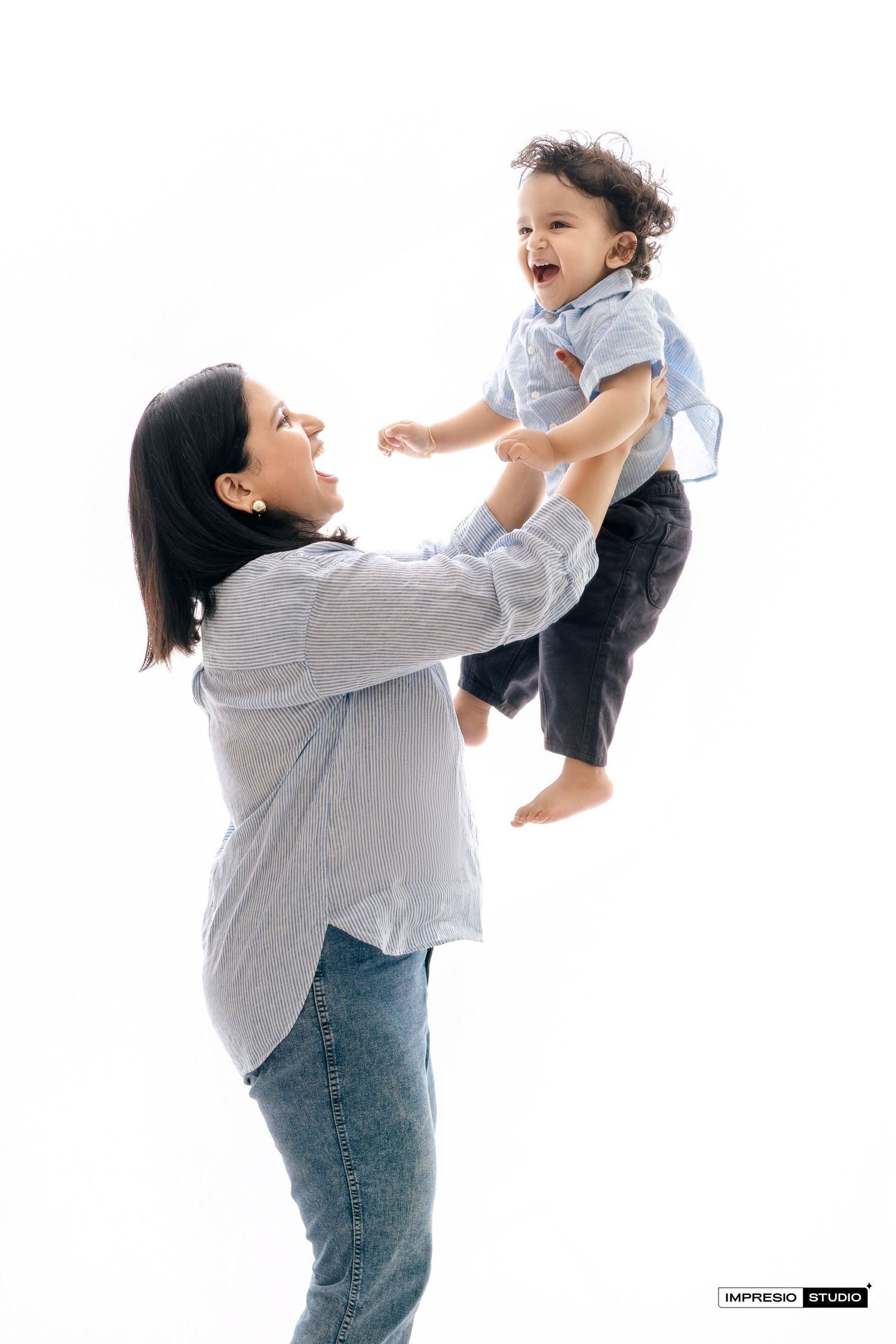 A caregiver in grey shirt and jeans lifts a joyful young child wearing a grey shirt and black pants against white background.