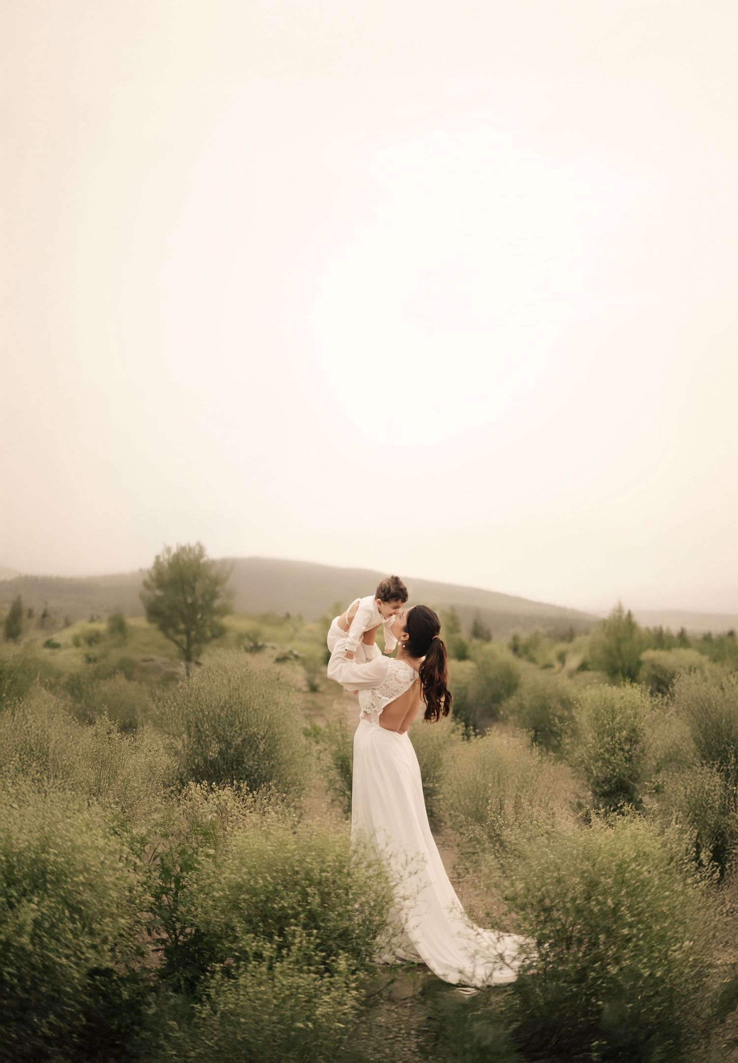 A figure in a flowing white wedding gown lifts a small child while standing among wild grasses on a hillside.