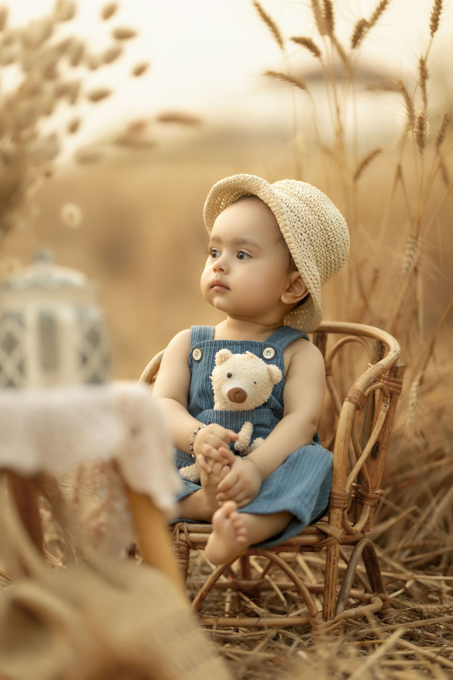 Sweet toddler in denim overalls and straw hat sits on wicker chair amidst tall wheat grass in warm sepia tones.