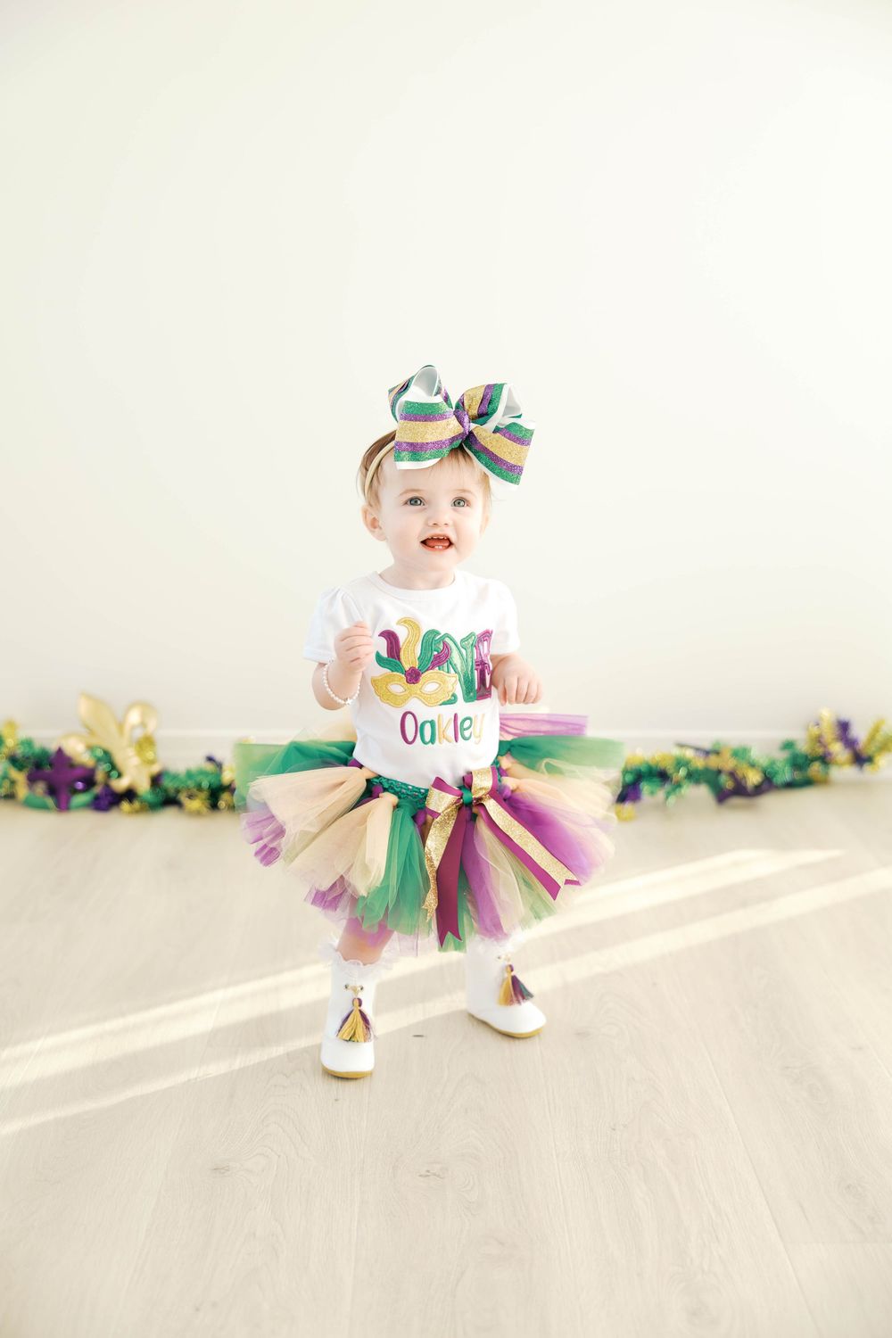 A baby in a colorful tutu dress and bow crawls and plays on a light colored floor with festive garland in the background.