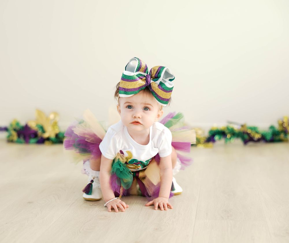 Baby in colorful tutu and headband crawls across white floor while paper garland decorates the background.