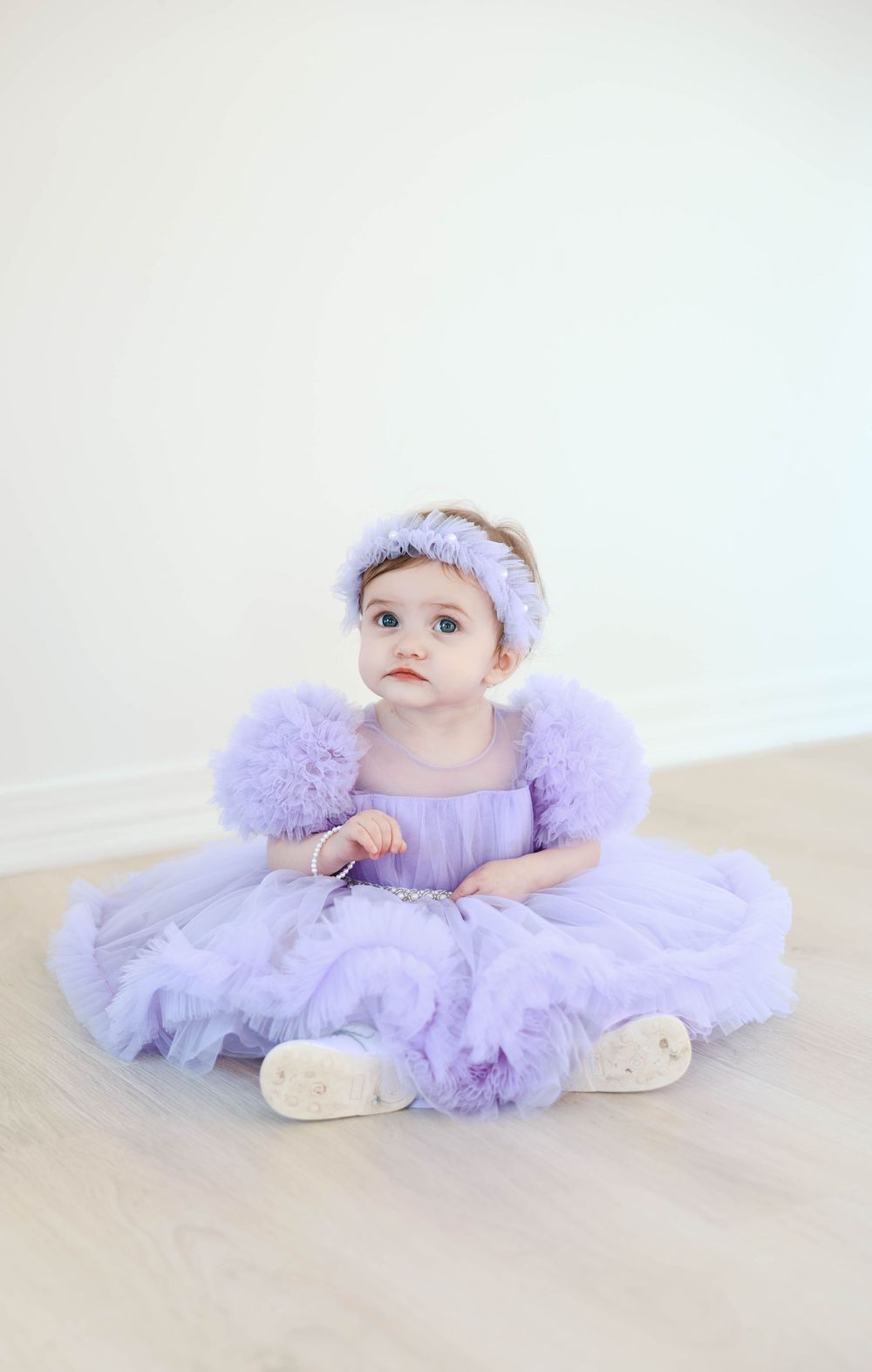 Baby wearing lavender tulle dress and matching headband lies on light beige background in artistic portrait series.