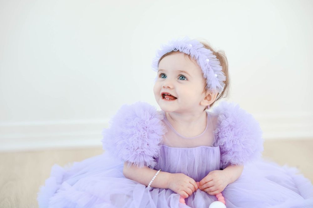 A baby wearing a lavender tulle dress and matching headband sits on a light colored floor.