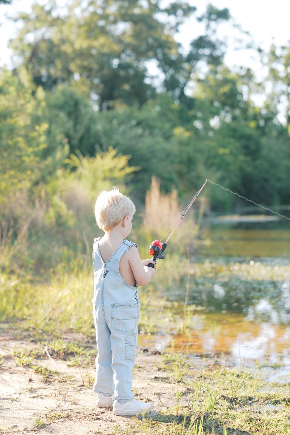 Toddler boy in denim overalls fishing with a rod by a sunny pond surrounded by green trees.