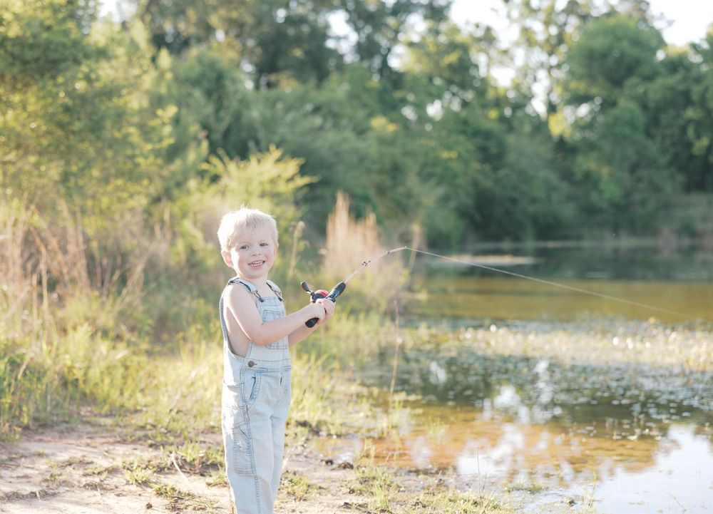 A smiling blonde toddler boy holds a fishing rod by a serene pond surrounded by lush green trees and tall grass.