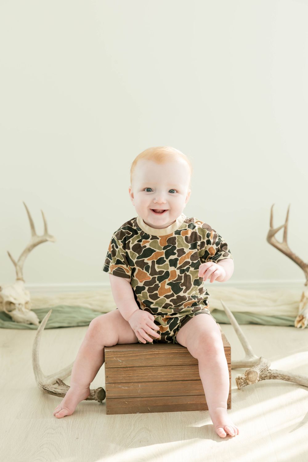 Smiling baby in camo onesie sitting on wooden box with deer antlers in background.