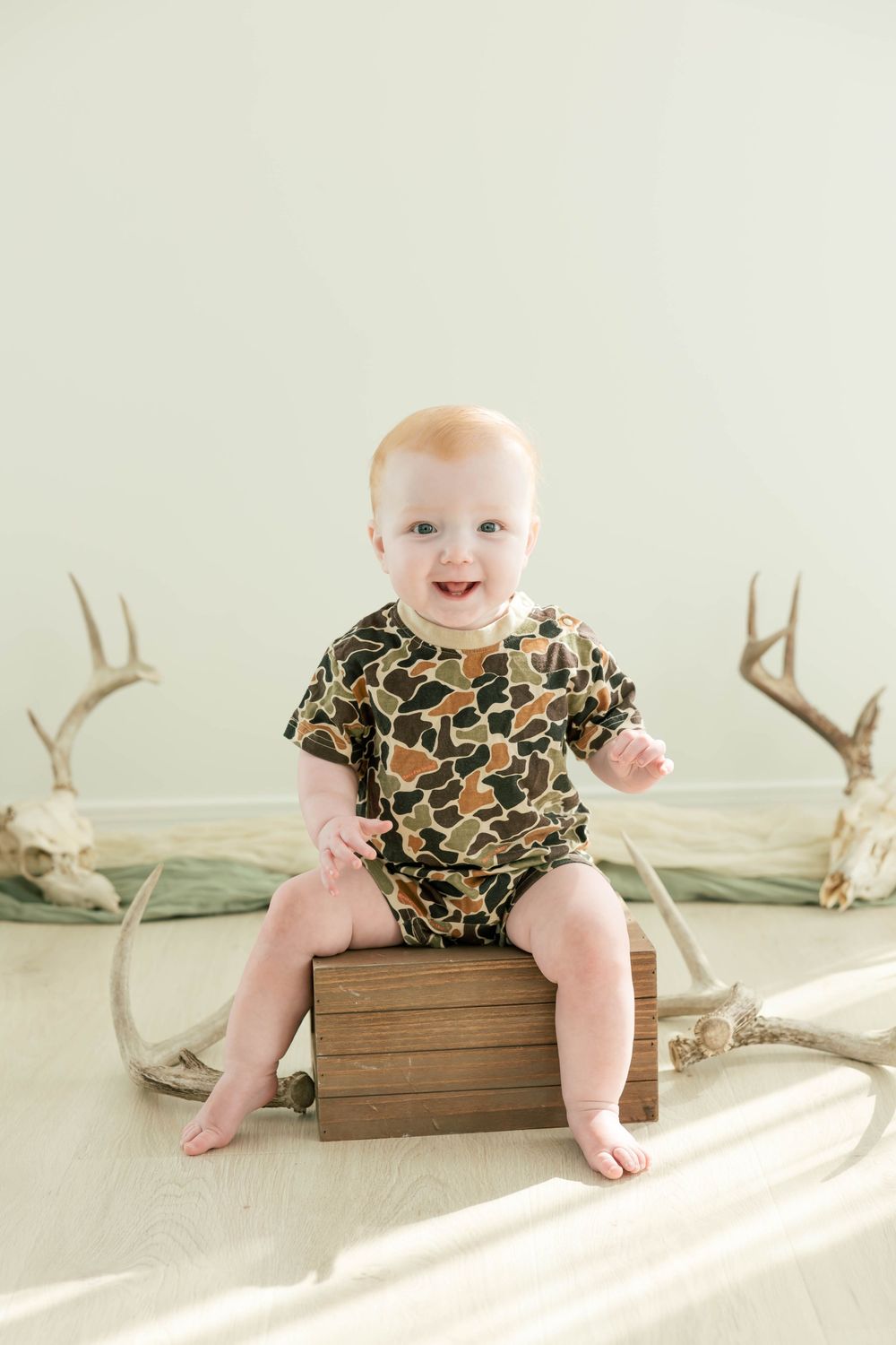Smiling baby in camo romper sitting on wooden crate surrounded by deer antlers on light green background.