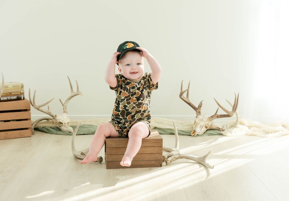 Baby in camo onesie and hat sits on wooden box surrounded by deer antlers in a bright studio photo.