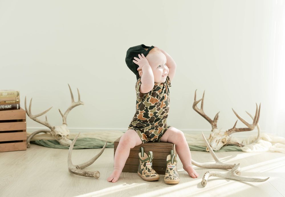 Baby in floral romper and cowboy hat sits on vintage suitcase surrounded by deer antlers in rustic photo shoot.