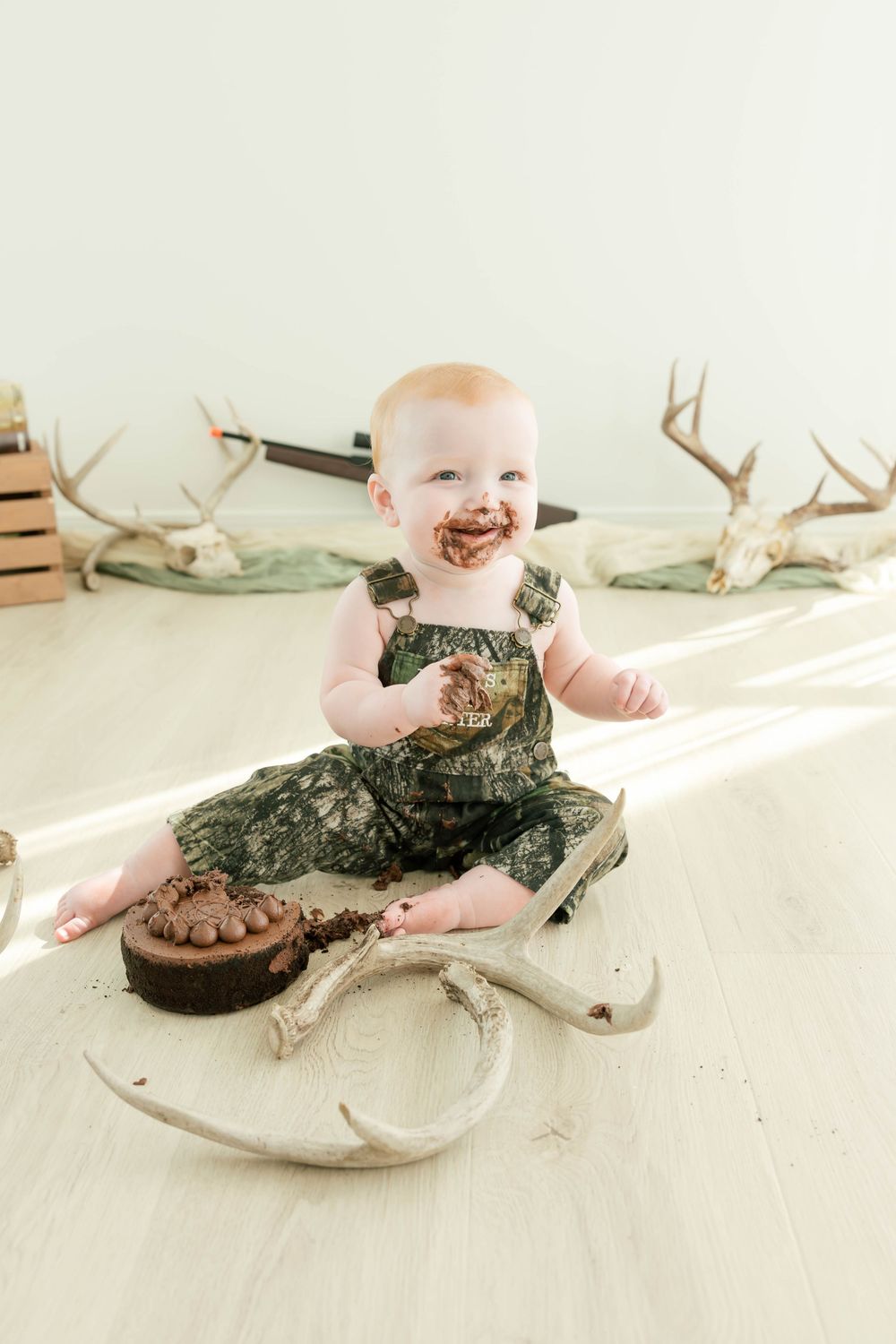Baby in camo outfit enjoys chocolate cake smash surrounded by deer antlers in bright white studio setting.