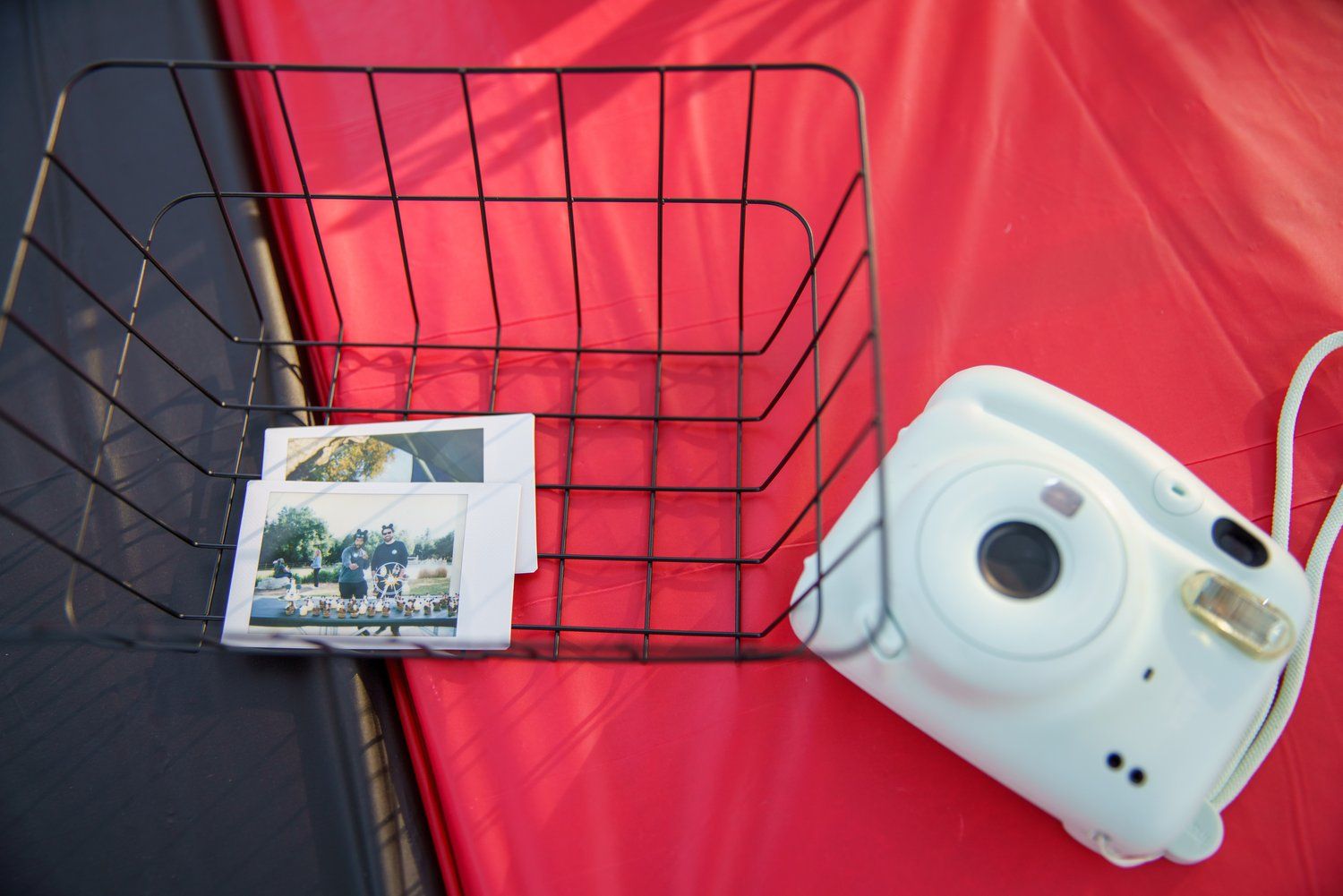 White Fujifilm Instax mini camera beside a wire basket with photos on a red background.