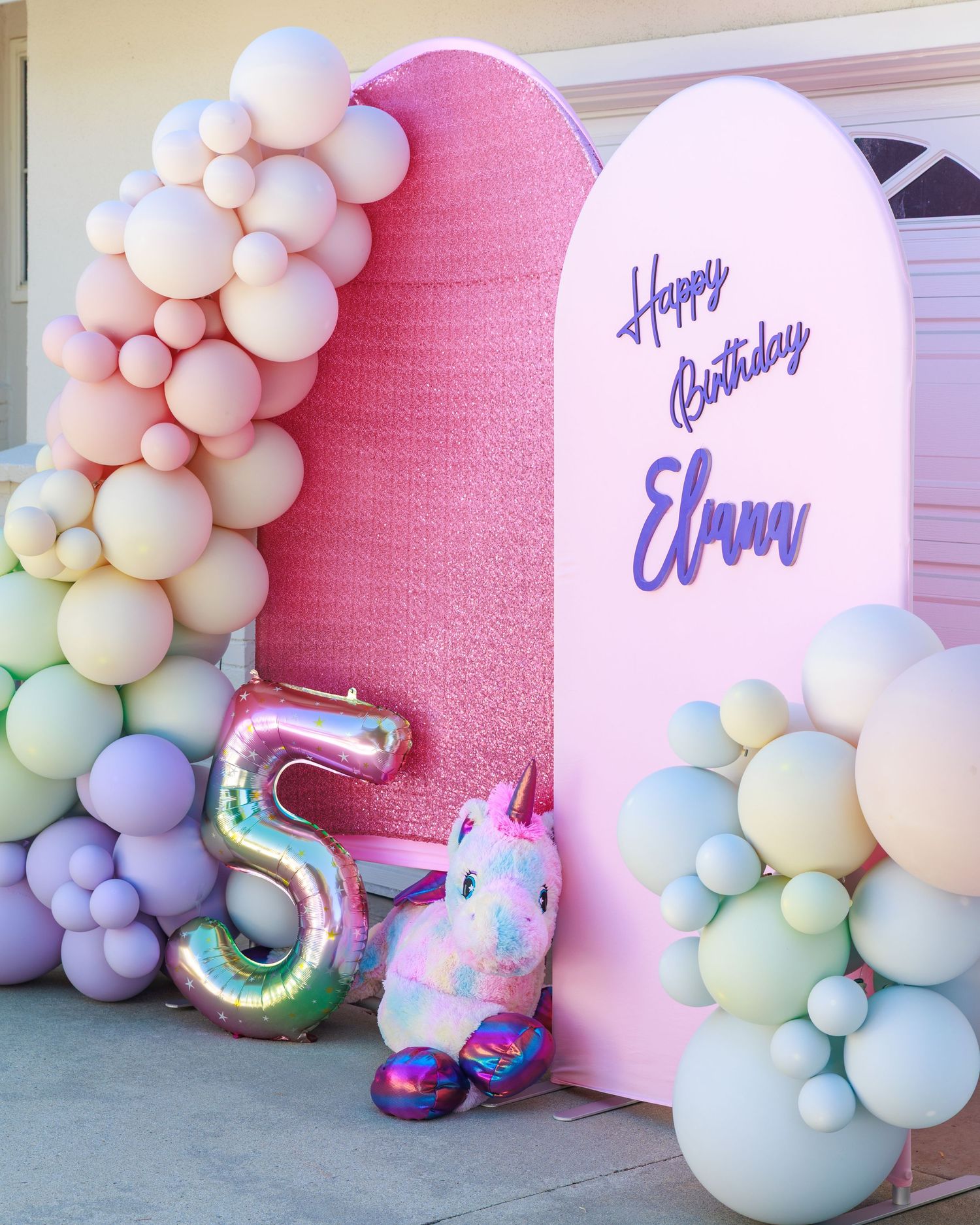 Pastel rainbow balloon garland with number 5 balloon and unicorn plush beside a pink Happy Birthday Elma backdrop.
