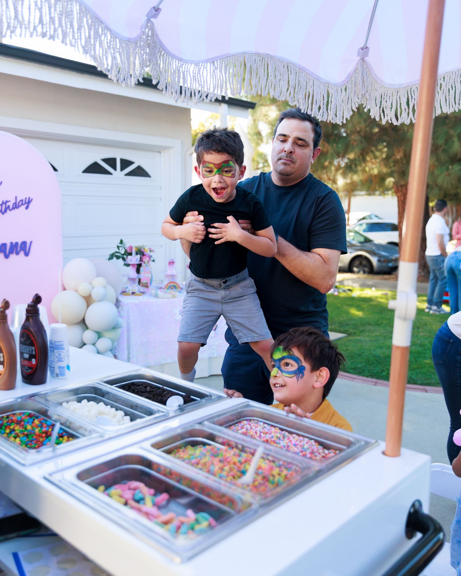 A man lifts a boy with face paint at a birthday party ice cream topping bar outdoors.