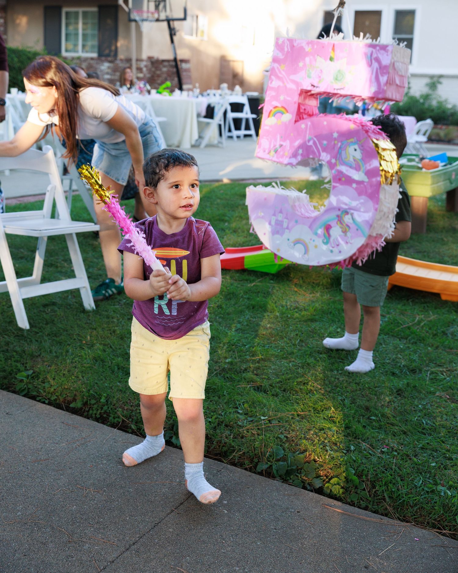 Young boy holding a stick next to a number 5 pinata at an outdoor birthday party celebration.