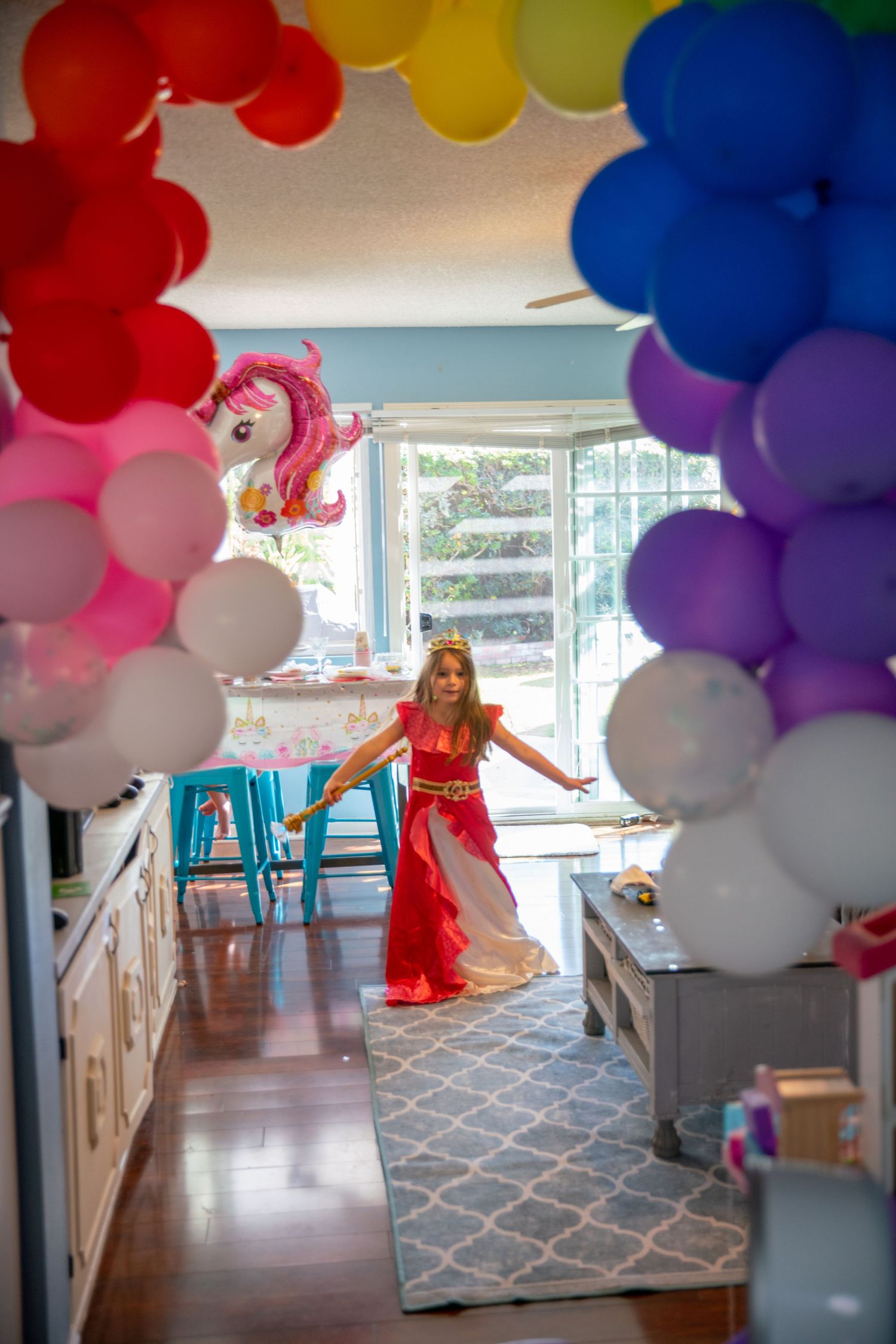 Girl in red princess costume poses under rainbow balloon arch at birthday party with unicorn balloon.