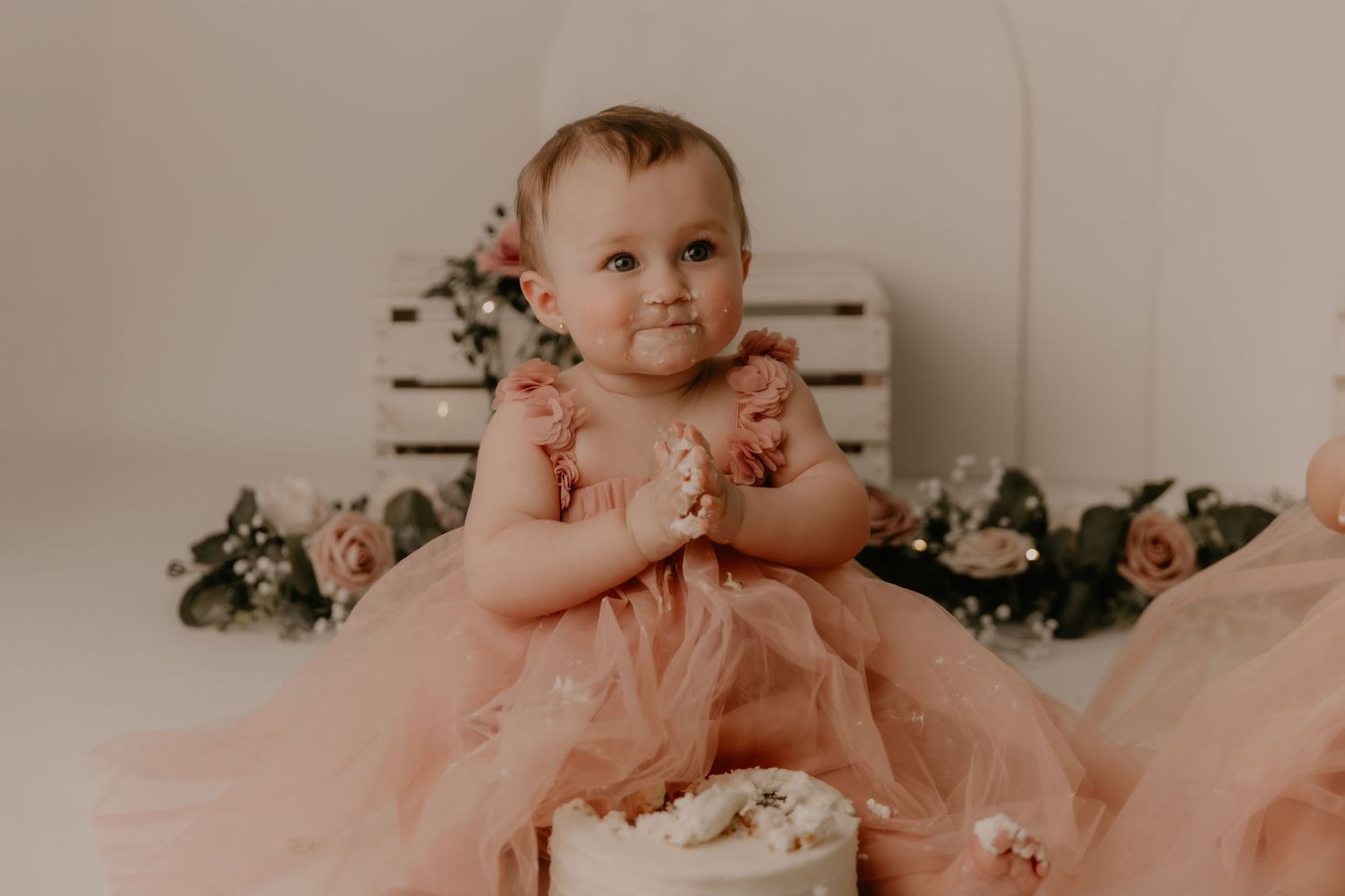 A small child in striped outfit sits on floor with white buttercream cake surrounded by floral decorations and holiday details.