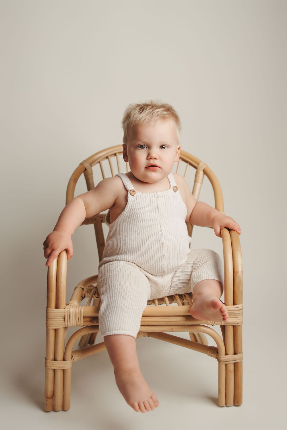 Baby wearing white ribbed outfit sits in natural rattan chair against neutral background.