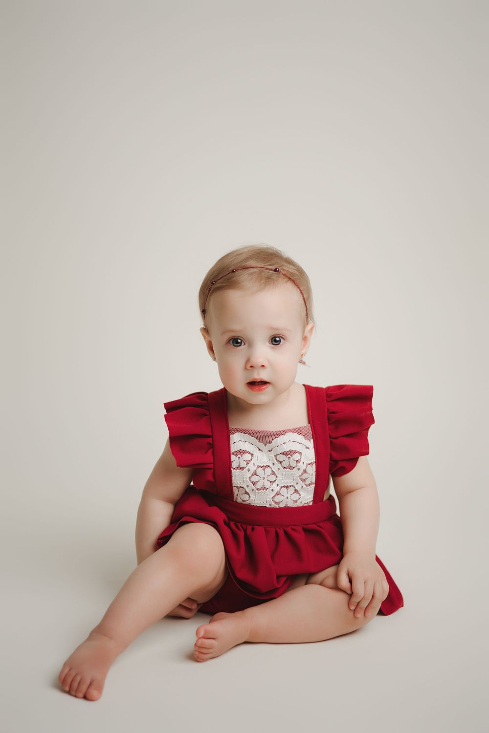 A small child sits in a red ruffled dress with white lace detail against a light gray background.