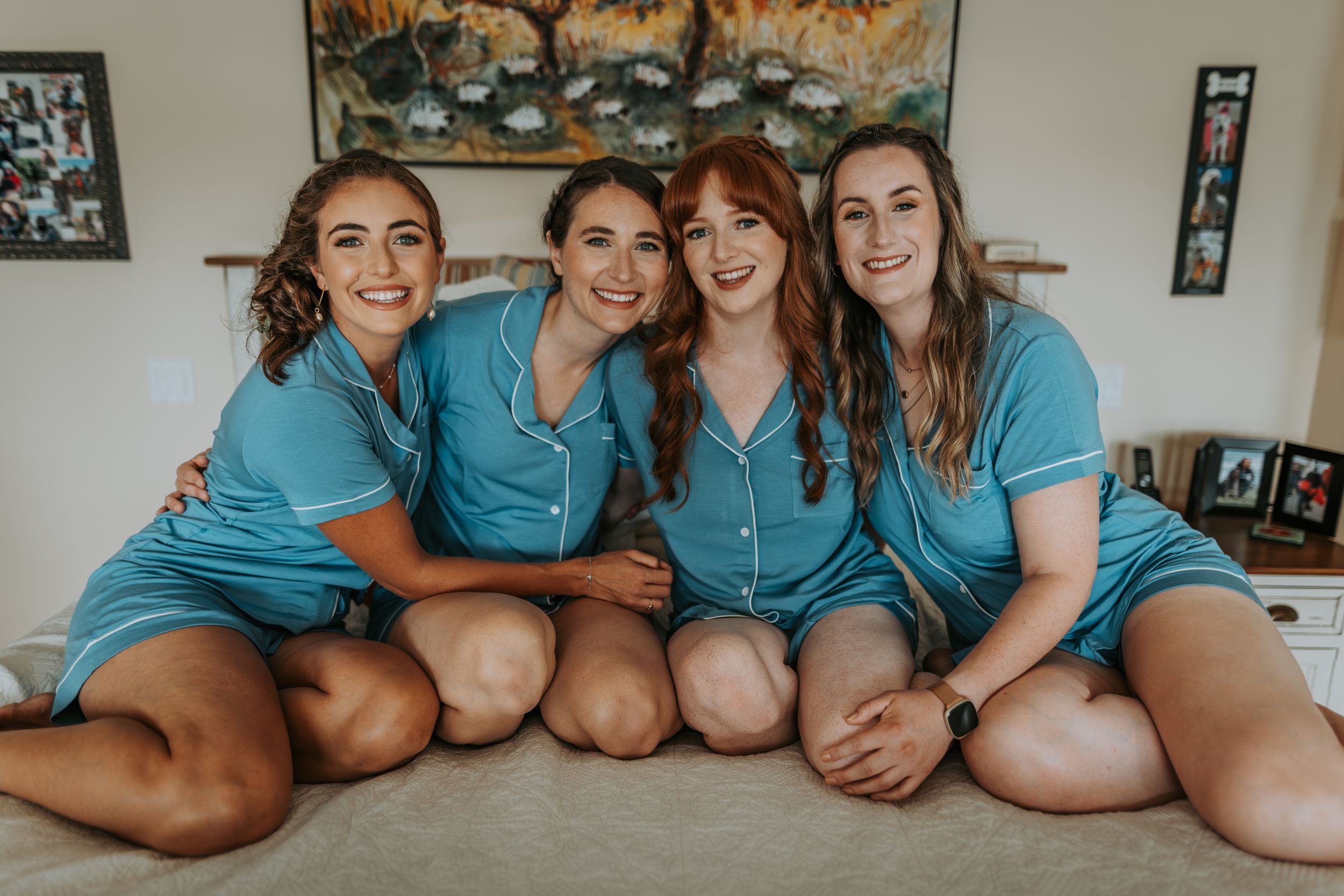 Four bridesmaids in matching teal satin pajamas smile together on a bed.