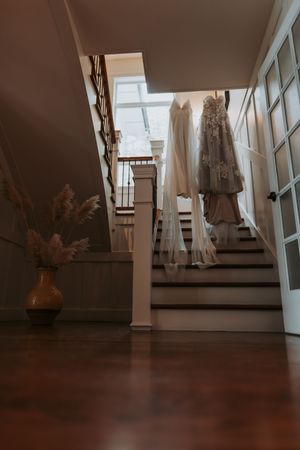 Two elegant wedding dresses hang on a staircase railing in a softly lit home interior.