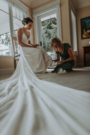 A bride in a long white gown has her shoes fastened by a helper in a bright room.