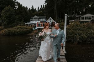 A bride and companion walk hand in hand along a wooden dock beside a lake.