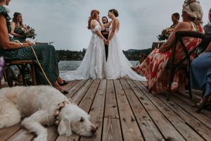 Two brides exchange vows on a lakeside dock as guests watch and a white dog rests nearby.