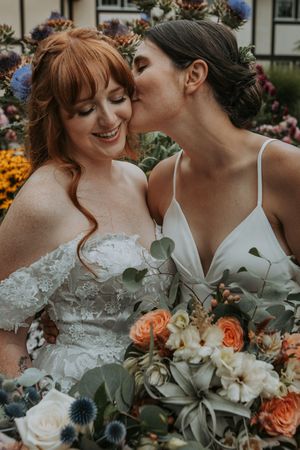 Two brides share a tender kiss surrounded by colorful garden flowers and lush bouquets.