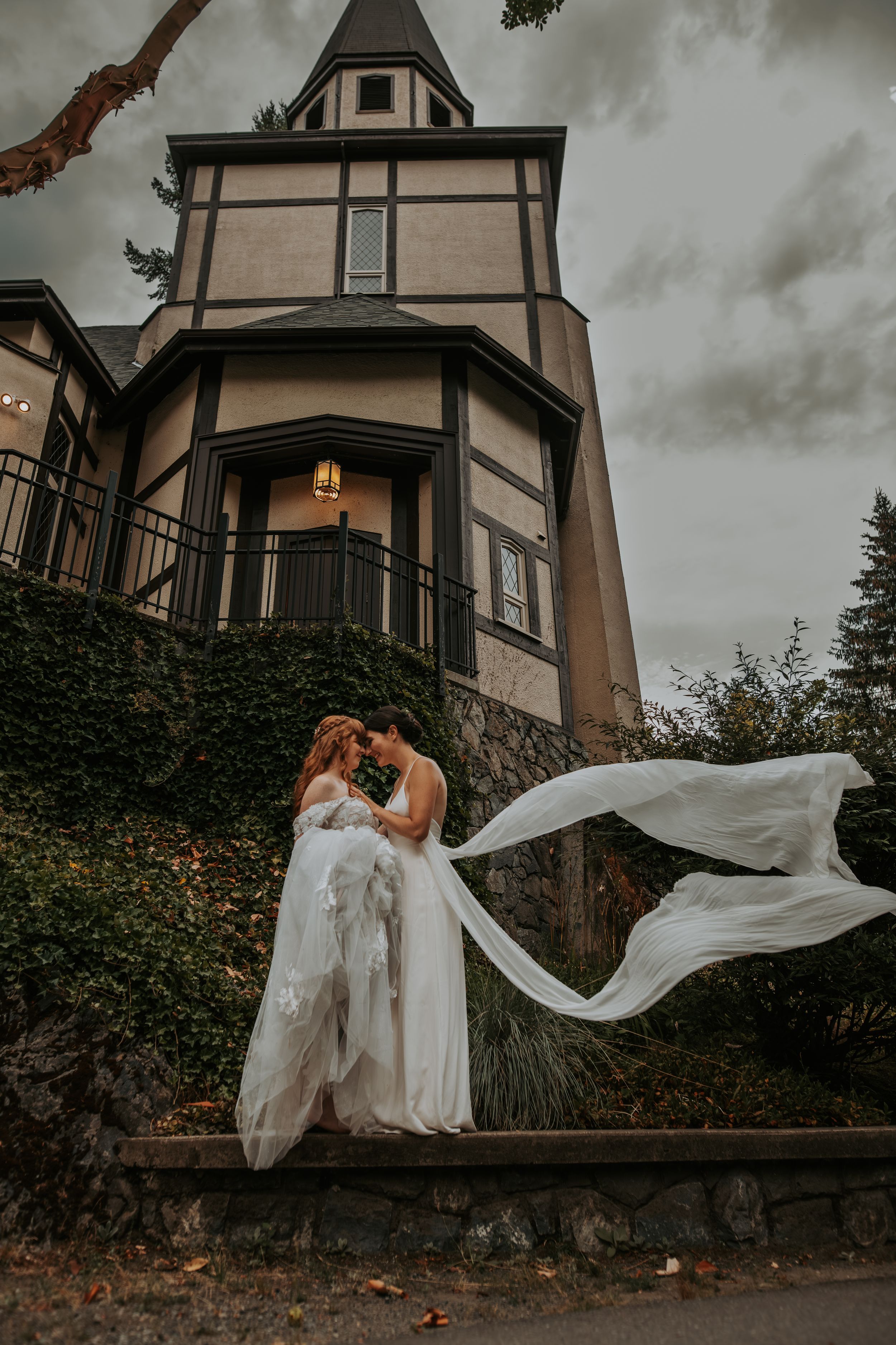 Couple embraces in front of a Tudor-style mansion with veil flowing in the wind.