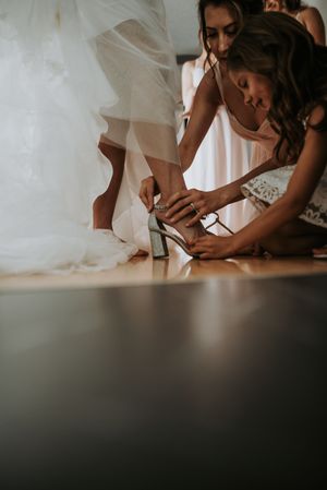 Bridesmaids helping bride put on elegant heeled wedding shoes before ceremony.