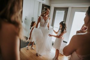 Bride in lace wedding gown surrounded by flower girls in bright room.