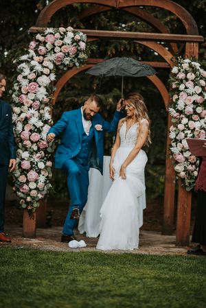Groom in blue suit stomping grapes with bride under umbrella at floral arch.
