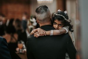 Young flower girl in a crown hugs a man in a black suit at a wedding.