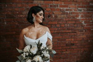 Bride with vitiligo in an off-shoulder gown holds white bouquet against a brick wall.