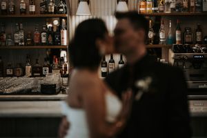 Bride and groom sharing a romantic kiss at a bar with bottle-lined shelves.